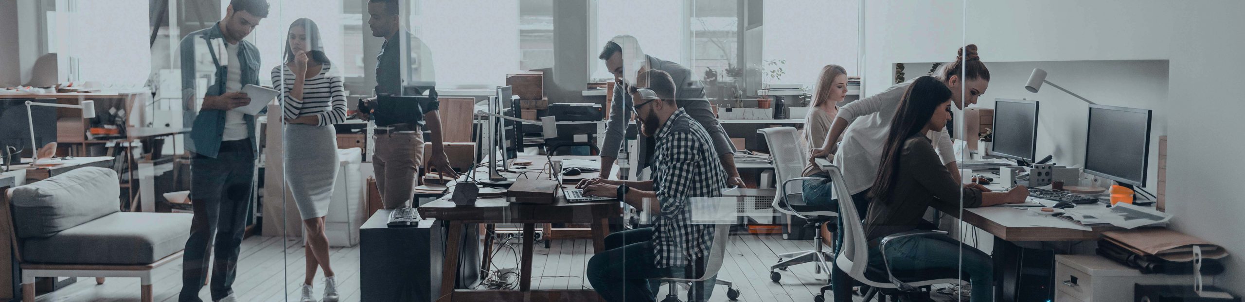 People working in an open office with multiple tables and computer monitors, some people conversing.