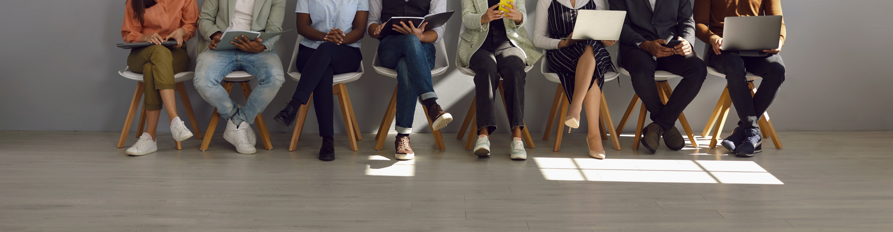 Wood floor and wall. Against the wall are 8 chairs. Only the legs and torso of the people sitting in the chairs are visible.