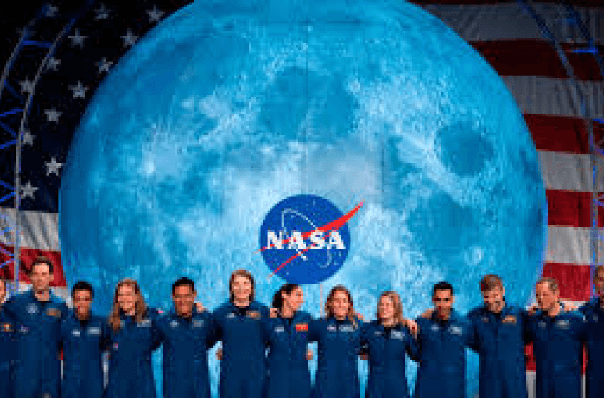 A line of over 12 astronauts standing in front of a very large screen depicting the US flag overlaid with an image of the Moon, which has the NASA logo at its center.