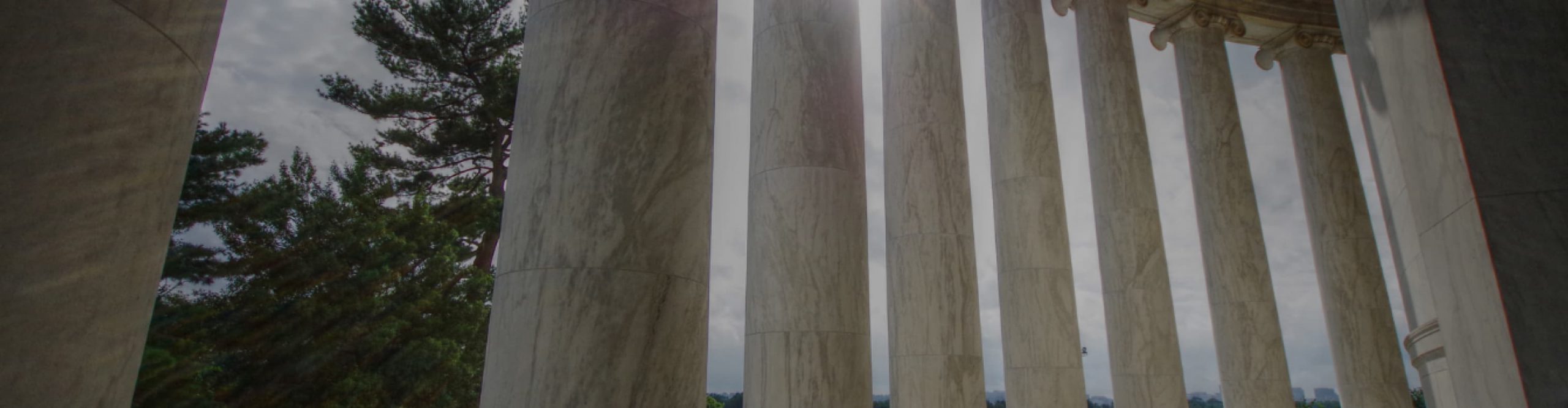 Marble columns foregrounded against trees and a cloudy sky.