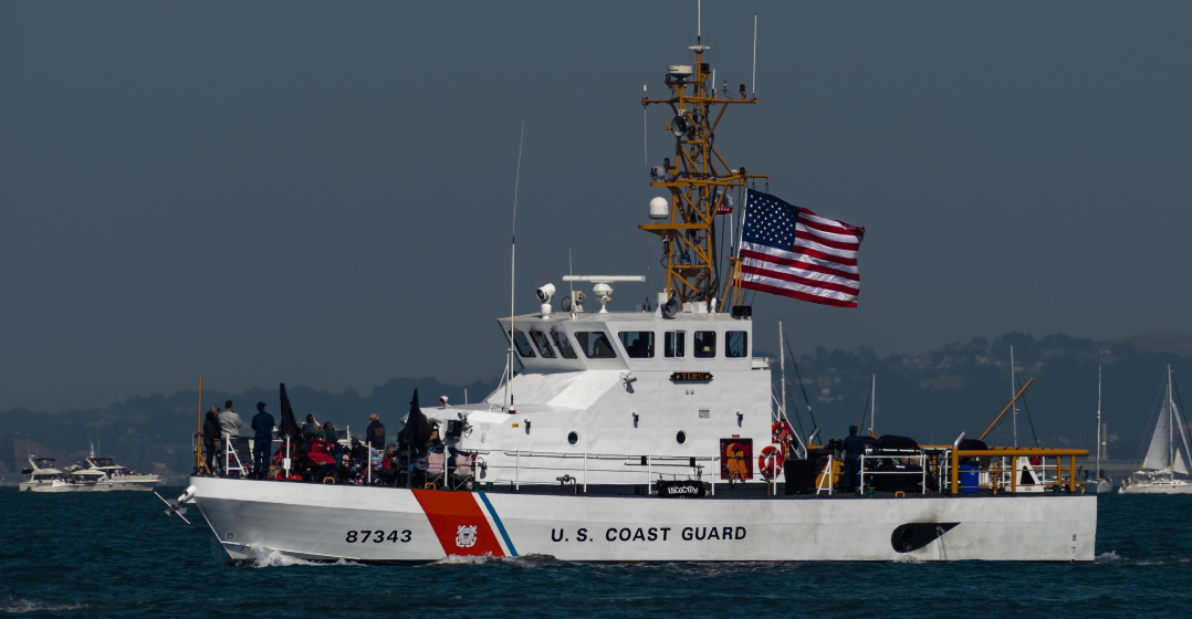 US Coast Guard vessel at sea with sailboats and low hills in the background.