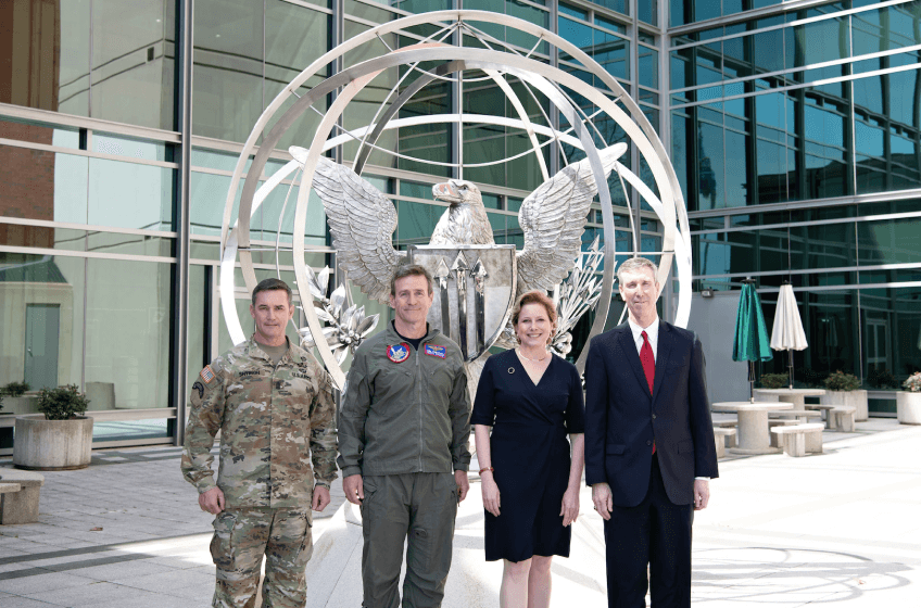 4 people stand in front of a building. Directly behind them is a large metal sphere with an eagle emblem on it. 2 are wearing military unforms, and 2 are wearing business attire.