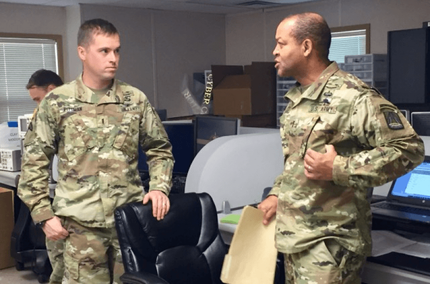 Two soldiers in camouflage uniforms stand talking to each other in an office setting.