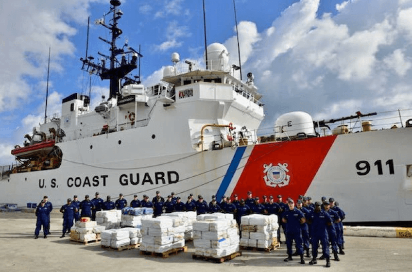 US Coast Guard vessel docked. In front of it stand about 50 people dressed in their blue Coast Guard uniforms and caps.