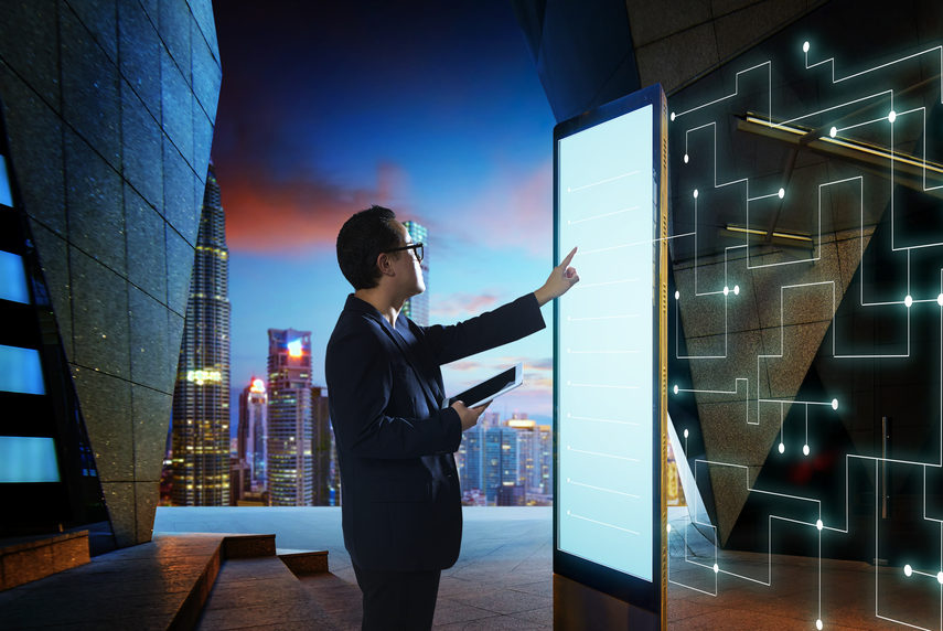 Man in business attire touching a very tall touch screen device. A cityscape visible in the background.