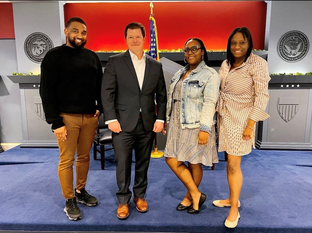 FedWriters employees pose together. Behind the group of four are the American flag and the seal of the Department of the Navy.