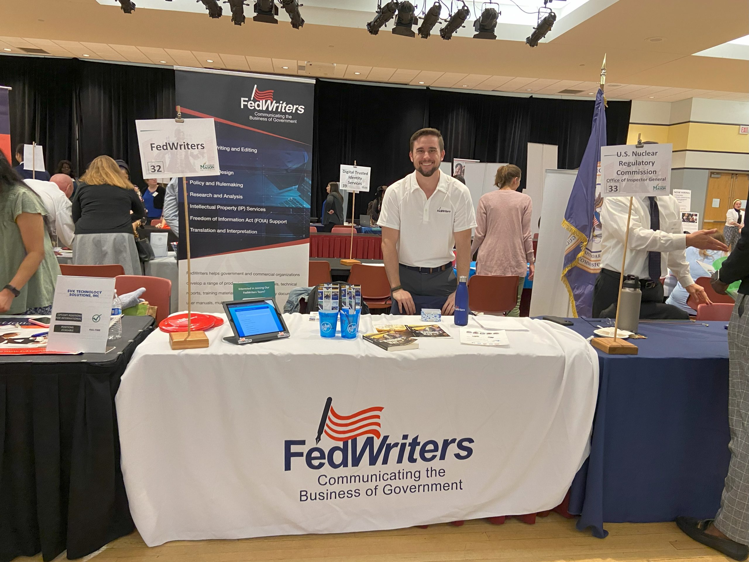 FedWriters' booth at a recruiting fair. Table covered in a white cloth with the FedWriters logo has cups and samples of FedWriters' publications.
